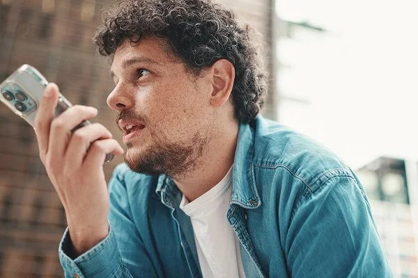 Junger, dunkelhaariger Mann mit Bart im Jeanshemd, der sitzt und in sein Smartphone spricht. Im Hintergrund eine moderne Stadtkulisse.
