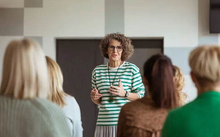 In einem Veranstaltungsraum steht eine Frau einer Gruppe von mehreren anderen Frauen gegenüber. Sie spricht zu den Frauen, die nur von hinten zu sehen sind.