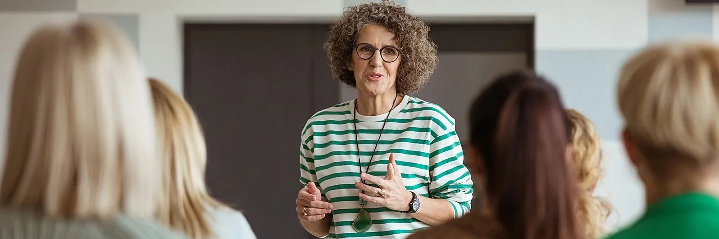 In einem Veranstaltungsraum steht eine Frau einer Gruppe von mehreren anderen Frauen gegenüber. Sie spricht zu den Frauen, die nur von hinten zu sehen sind.