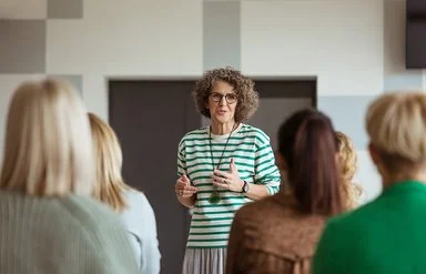 In einem Veranstaltungsraum steht eine Frau einer Gruppe von mehreren anderen Frauen gegenüber. Sie spricht zu den Frauen, die nur von hinten zu sehen sind.