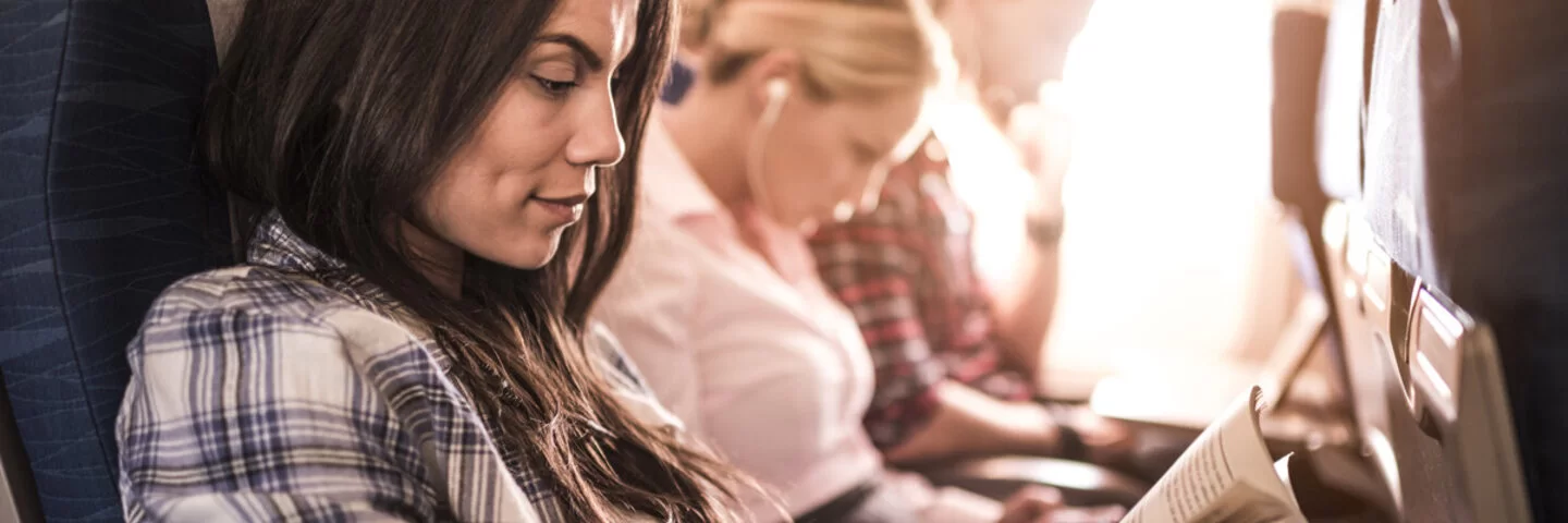Eine Frau sitzt neben zwei Mitreisenden im Flugzeug und liest ein Buch