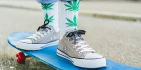 Im Bildausschnitt sind die Beine eines Menschen zu sehen. Er trägt Blue Jeans und graue Sneaker. Auf den weißen Socken sind grüne Marihuana-Pflanzen zu sehen. Er steht auf einem blauen Skateboard und fährt eine Straße entlang.