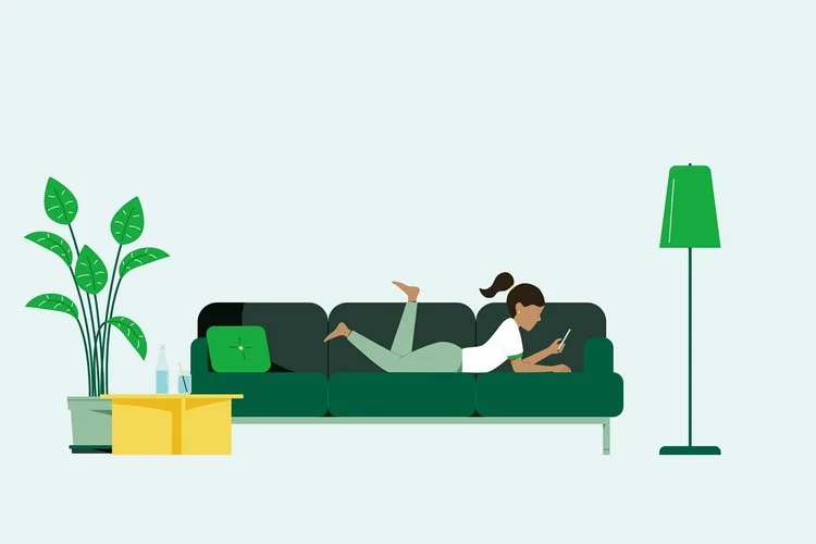 Frau liegt auf dem Bauch auf grünem Sofa und schaut auf ihr Smartphone, daneben Pflanze, gelber Tisch und grüne Stehlampe.