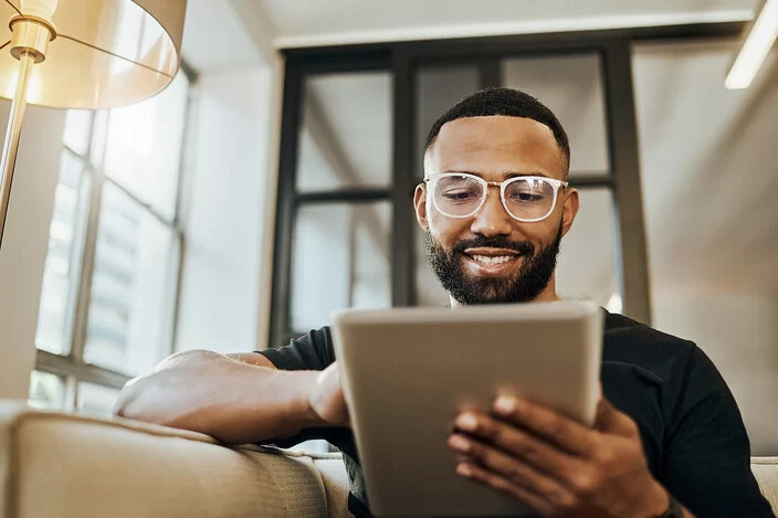 Ein junger Mann sitzt auf einem Sofa und schaut auf ein Tablet, das er in seinen Händen hält.