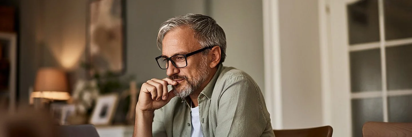 Ein älterer Mann mit Brille und grauem Haar sitzt nachdenklich vor einem Laptop an einem Esstisch.