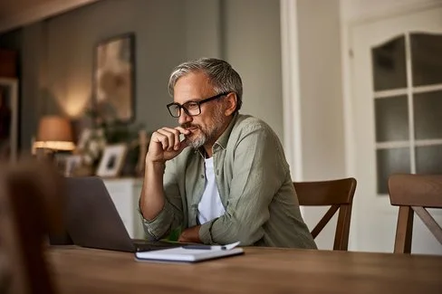 Ein älterer Mann mit Brille und grauem Haar sitzt nachdenklich vor einem Laptop an einem Esstisch.