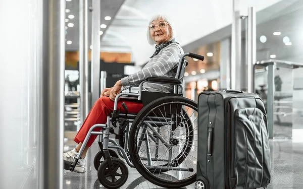 Eine Senorin sitzt im Rollstuhl und lächelt, neben ihr steht ein grauer Koffer.