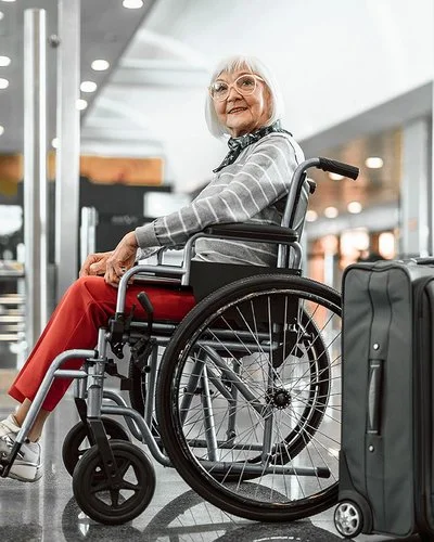 Eine Senorin sitzt im Rollstuhl und lächelt, neben ihr steht ein grauer Koffer.