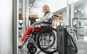 Eine Senorin sitzt im Rollstuhl und lächelt, neben ihr steht ein grauer Koffer.