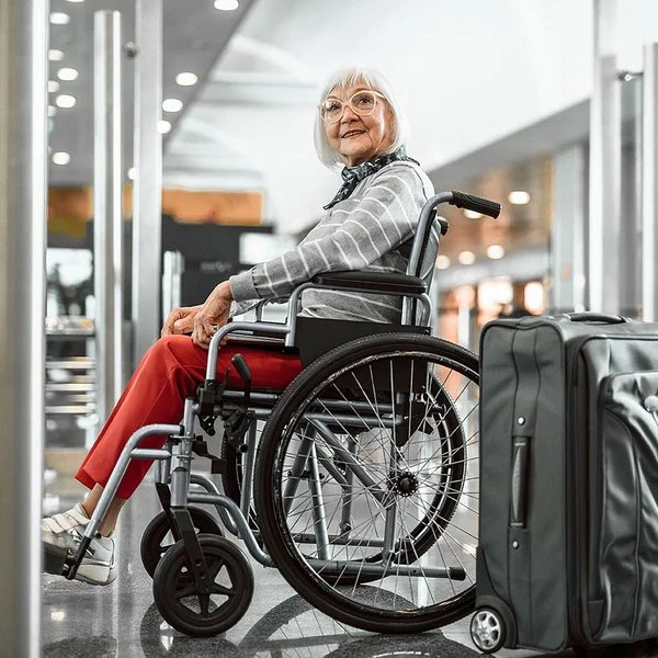 Eine Senorin sitzt im Rollstuhl und lächelt, neben ihr steht ein grauer Koffer.