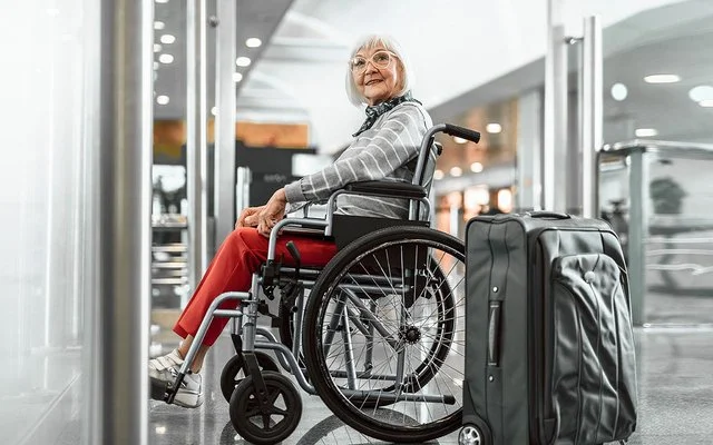 Eine Senorin sitzt im Rollstuhl und lächelt, neben ihr steht ein grauer Koffer.