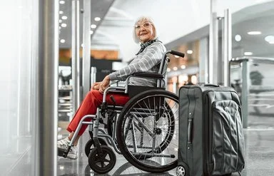 Eine Senorin sitzt im Rollstuhl und lächelt, neben ihr steht ein grauer Koffer.