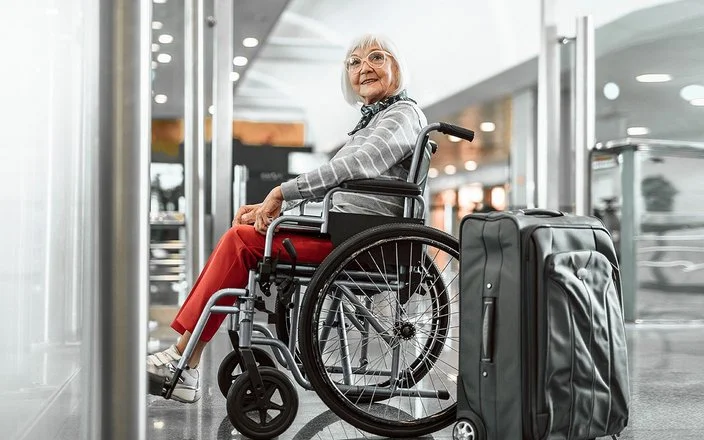 Eine Senorin sitzt im Rollstuhl und lächelt, neben ihr steht ein grauer Koffer.