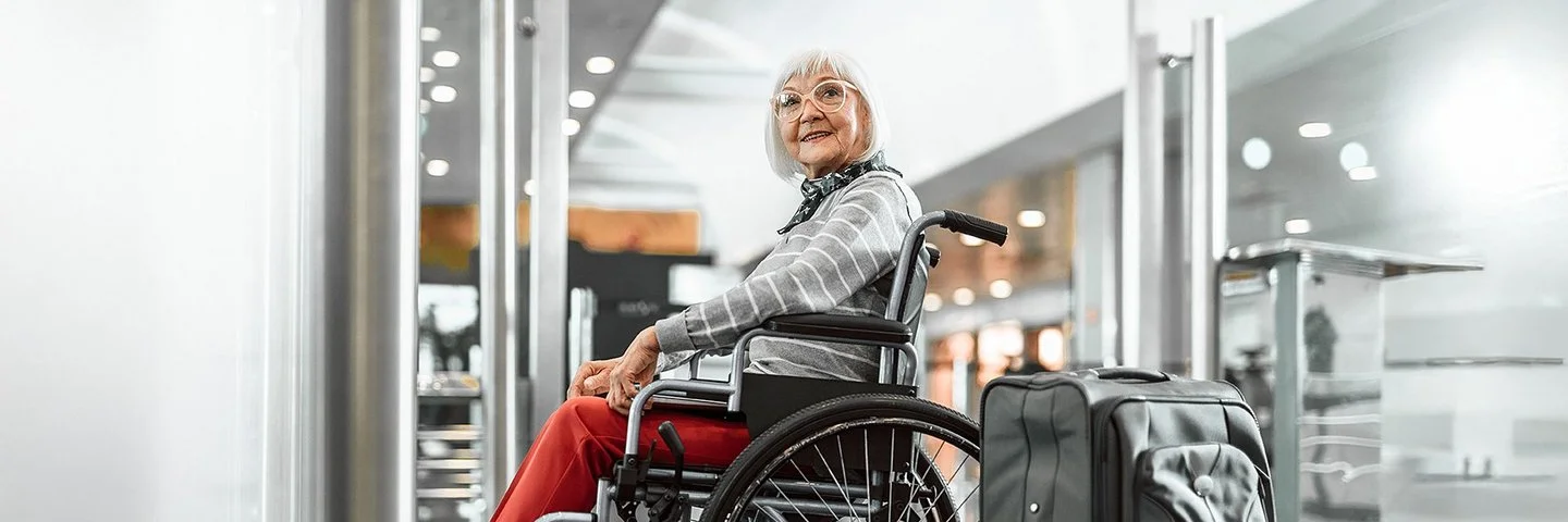 Eine Senorin sitzt im Rollstuhl und lächelt, neben ihr steht ein grauer Koffer.