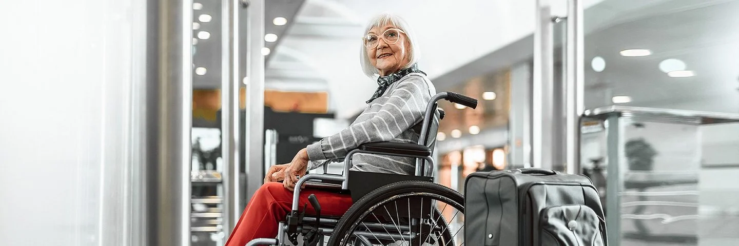 Eine Senorin sitzt im Rollstuhl und lächelt, neben ihr steht ein grauer Koffer.