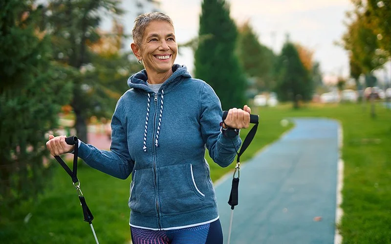 Eine ältere Frau trainiert mit einem Widerstandsband in einer Parkanlage.