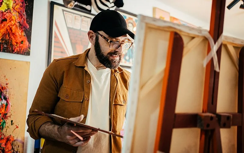 In einem Raum mit Bildern an der Wand steht ein Mann mit einer Palette in der rechten Hand vor einer Staffelei, auf der eine Leinwand steht. Er blickt auf die Leinwand, die nur schräg von hinten zu sehen ist und seine linke Hand verdeckt.