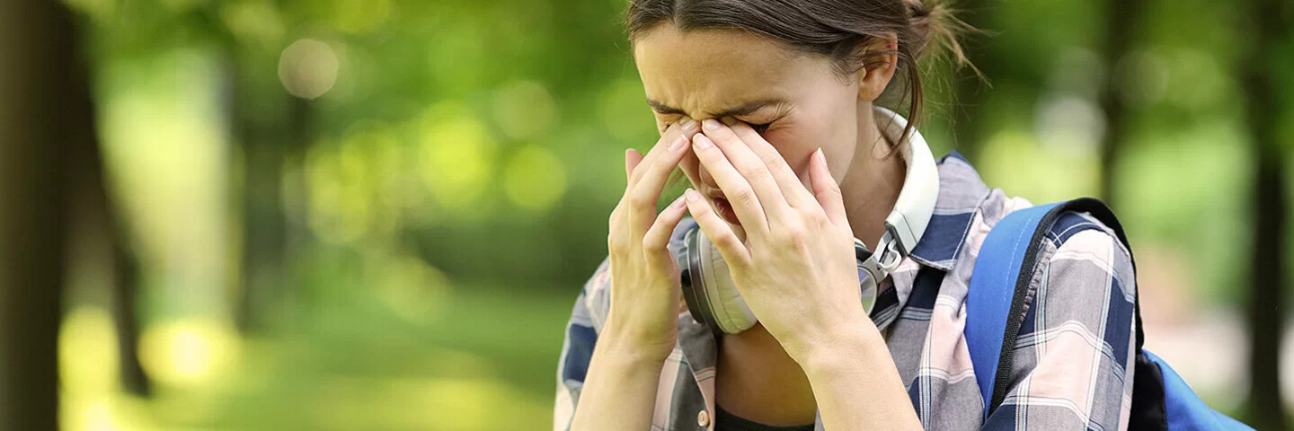 Eine junge Frau ist im Park unterwegs. Sie leidet an Allergiesymptomen und reibt sich die Augen.