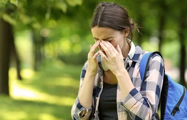 Eine junge Frau ist im Park unterwegs. Sie leidet an Allergiesymptomen und reibt sich die Augen.