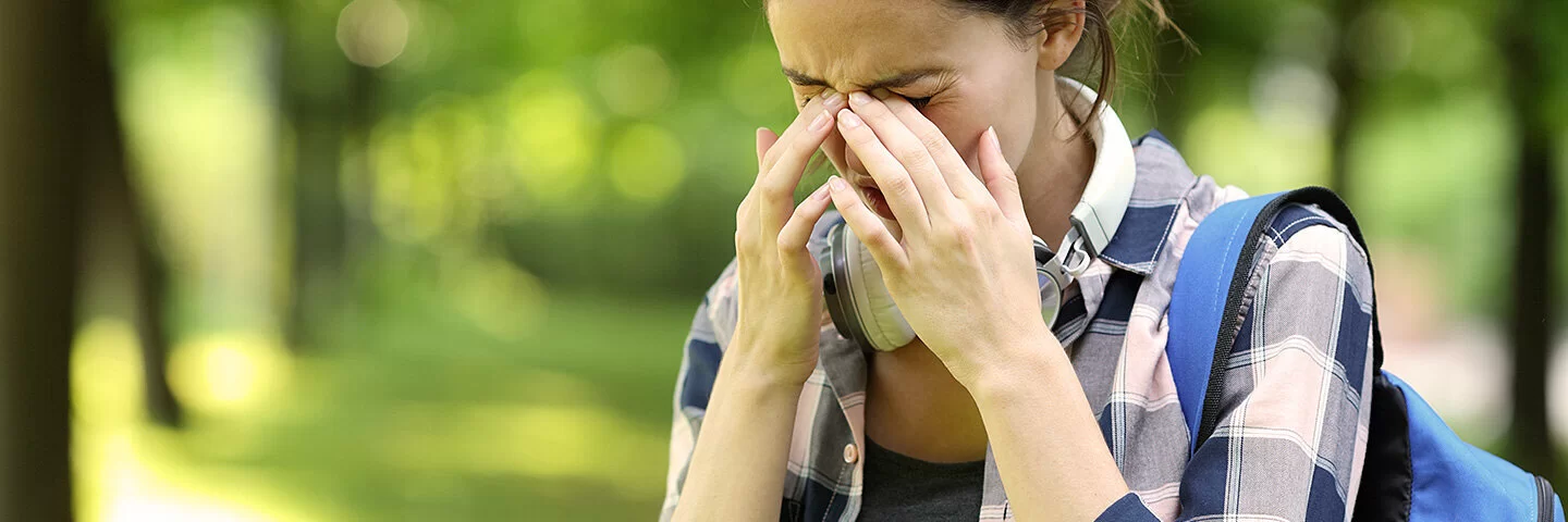 Eine junge Frau ist im Park unterwegs. Sie leidet an Allergiesymptomen und reibt sich die Augen.