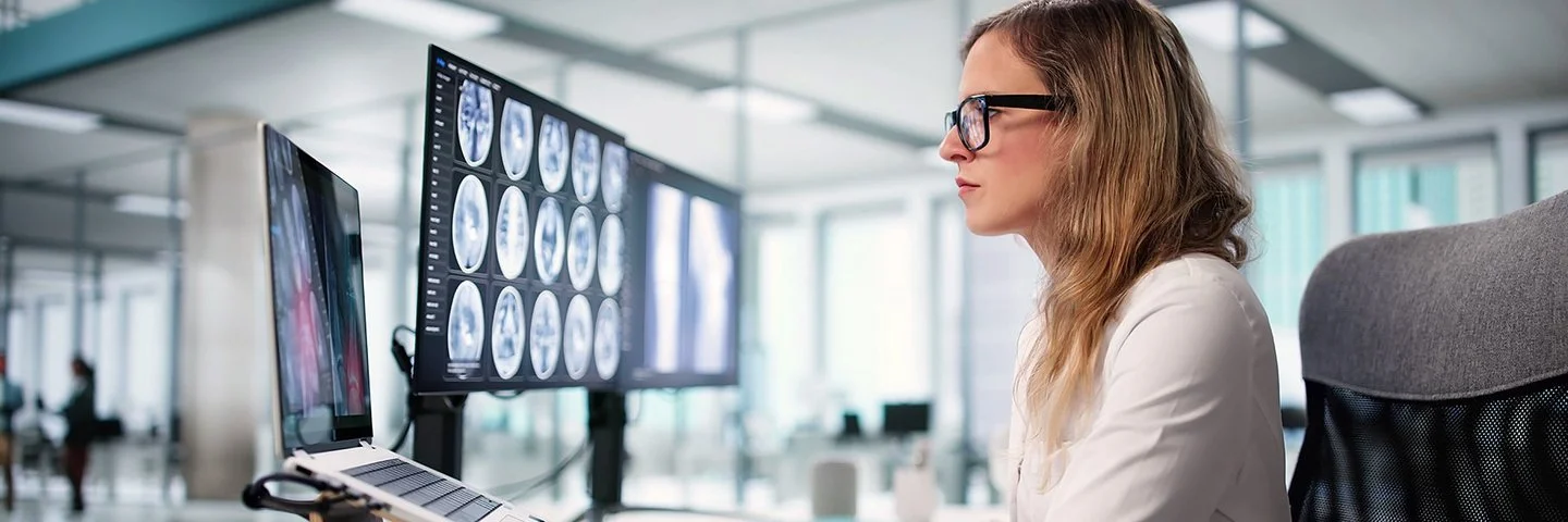 Eine Frau im weißen Kittel und mit schwarzer Brille sitzt an einem Schreibtisch in einer modernen Umgebung und betrachtet einen Gehirnscan auf einem Computer.