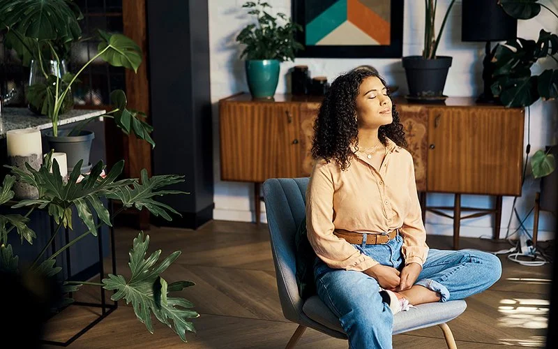 Eine Frau sitzt entspannt in einem Sessel, sie hat die Augen geschlossen und scheint zu meditieren. Im Hintergrund ein Wohnzimmer mit grünen Pflanzen.