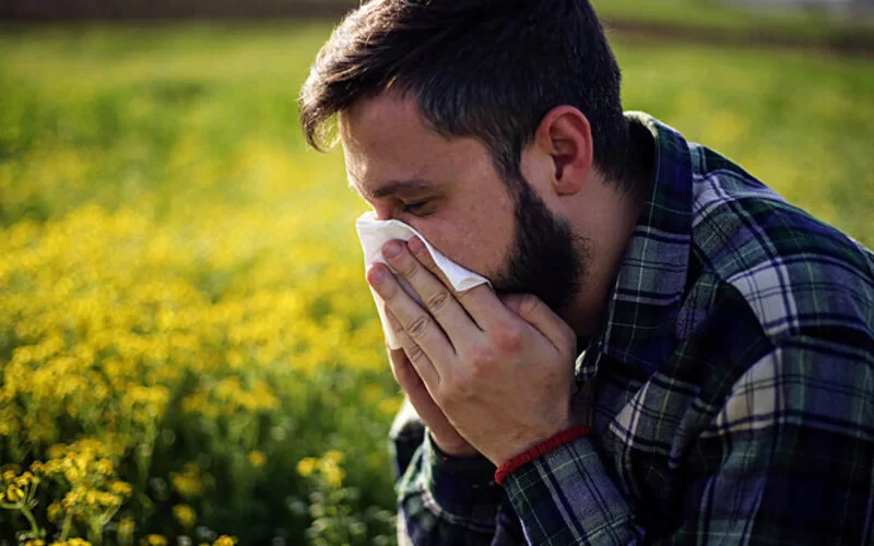 Ein Mann mit Allergie steht an einem Feld und niest in ein Taschentuch.