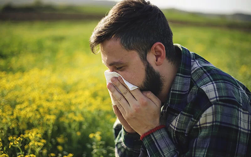 Ein Mann mit Allergie steht an einem Feld und niest in ein Taschentuch.