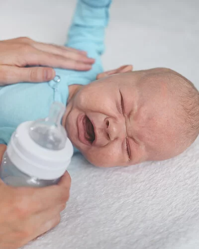 Ein Neugeborenes liegt auf dem Bett, kneift die Augen zusammen und schreit. Die Mutter hat die linke Hand auf die Brust des Kindes gelegt, um es zu trösten. In der rechten Hand hält sie eine Babyflasche.