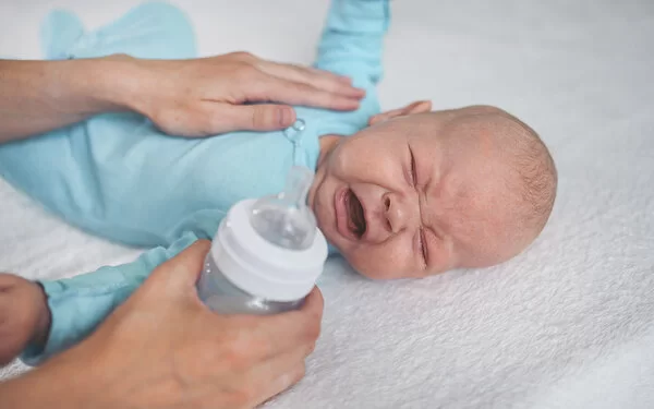 Ein Neugeborenes liegt auf dem Bett, kneift die Augen zusammen und schreit. Die Mutter hat die linke Hand auf die Brust des Kindes gelegt, um es zu trösten. In der rechten Hand hält sie eine Babyflasche.