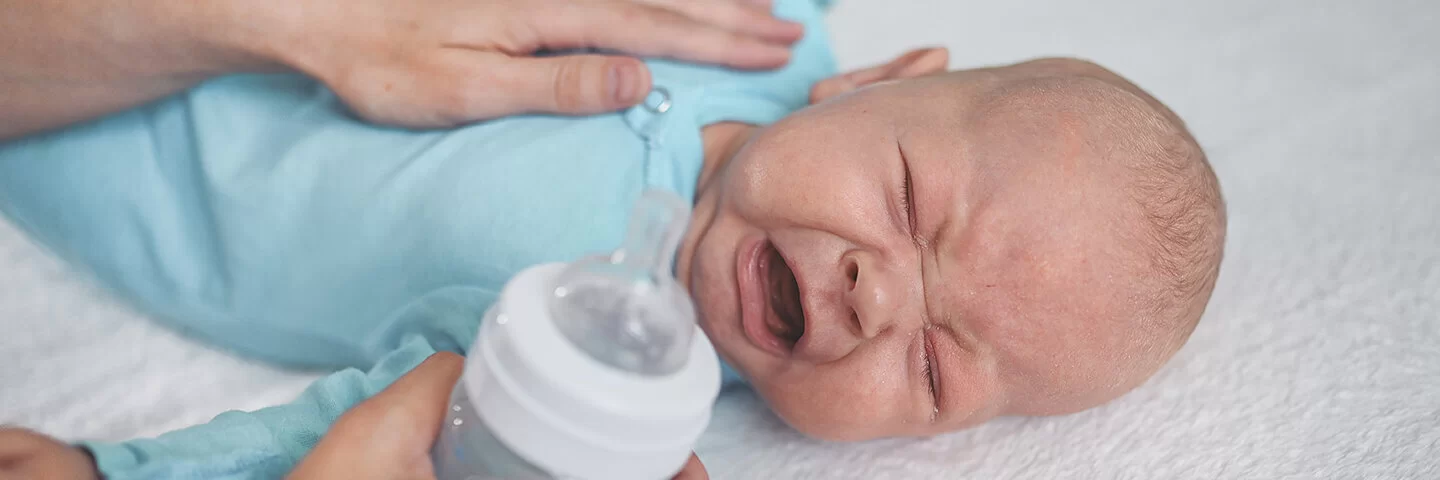 Ein Neugeborenes liegt auf dem Bett, kneift die Augen zusammen und schreit. Die Mutter hat die linke Hand auf die Brust des Kindes gelegt, um es zu trösten. In der rechten Hand hält sie eine Babyflasche.