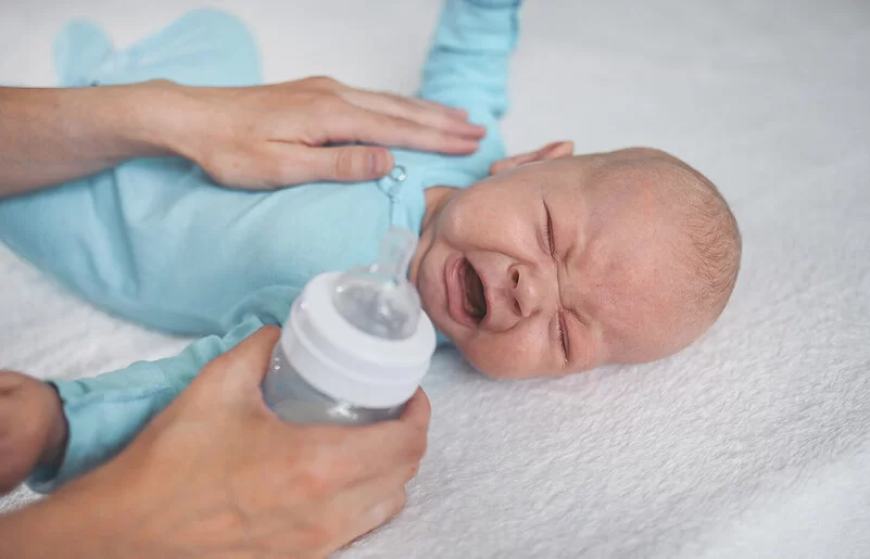 Ein Neugeborenes liegt auf dem Bett, kneift die Augen zusammen und schreit. Die Mutter hat die linke Hand auf die Brust des Kindes gelegt, um es zu trösten. In der rechten Hand hält sie eine Babyflasche.