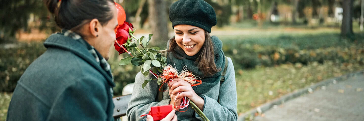 Ein junger Mann überhäuft eine junge Frau mit Präsenten: Blumen, Herzluftballons und eine kleine rote Geschenkschachtel.