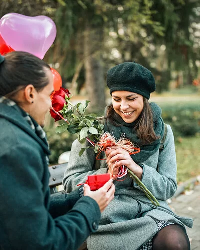 Ein junger Mann überhäuft eine junge Frau mit Präsenten: Blumen, Herzluftballons und eine kleine rote Geschenkschachtel.