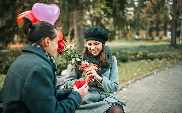 Ein junger Mann überhäuft eine junge Frau mit Präsenten: Blumen, Herzluftballons und eine kleine rote Geschenkschachtel.