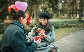 Ein junger Mann überhäuft eine junge Frau mit Präsenten: Blumen, Herzluftballons und eine kleine rote Geschenkschachtel.