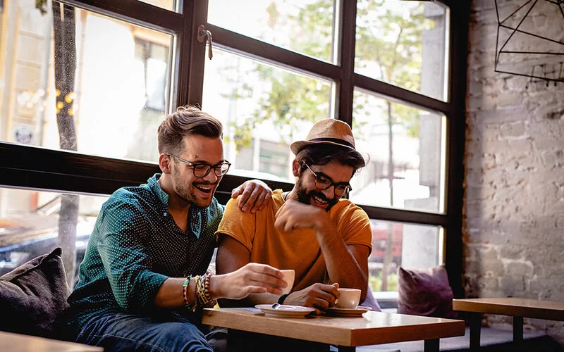Zwei junge Männer sitzen lachend am Tisch in einem Café. Auf dem Tisch stehen Kaffeetassen. Einer der beiden legt die Hand auf die Schulter des anderen.