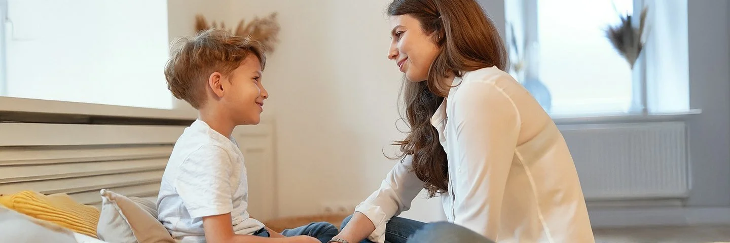 Eine junge Frau und ein junges Kind sitzen sich im Schneidersitz gegenüber und lächeln sich an.