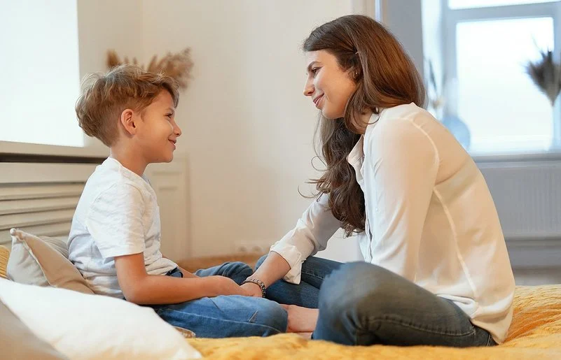 Eine junge Frau und ein junges Kind sitzen sich im Schneidersitz gegenüber und lächeln sich an.