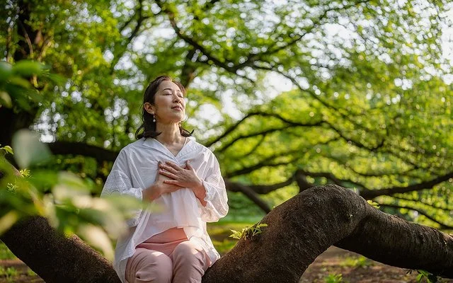 Eine junge Frau sitzt auf einem Baumstamm in der Natur und meditiert. Dabei hält sie sich die Hände ans Herz und hat die Augen geschlossen.