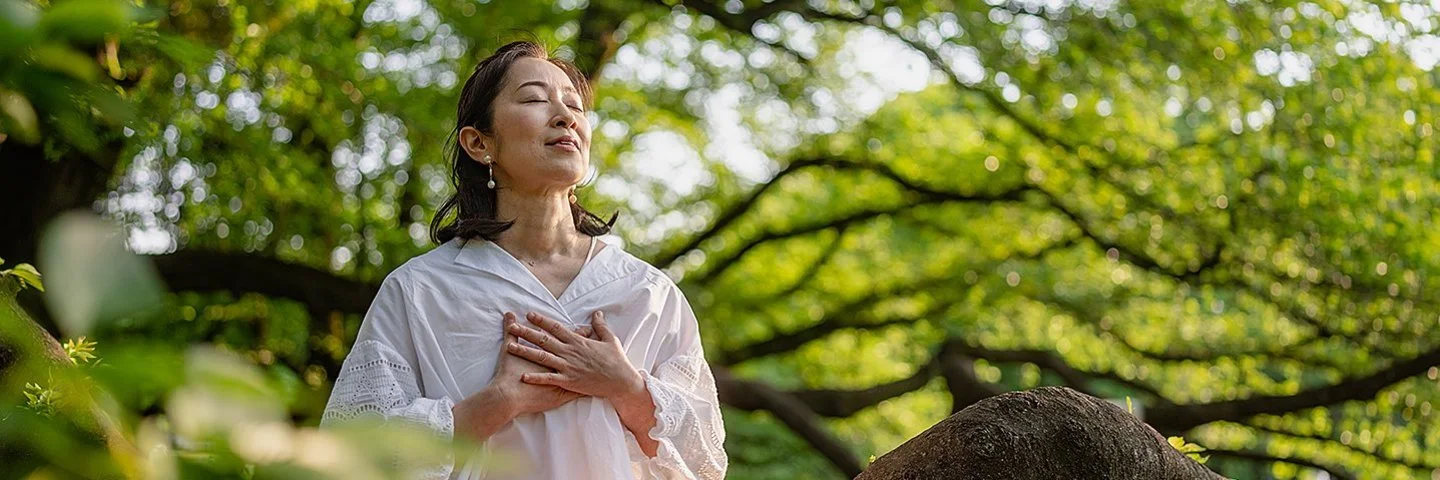 Eine junge Frau sitzt auf einem Baumstamm in der Natur und meditiert. Dabei hält sie sich die Hände ans Herz und hat die Augen geschlossen.