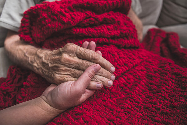 Eine Person hält die Hände einer sterbenden, alten Person mit einer roten Wolldecke.