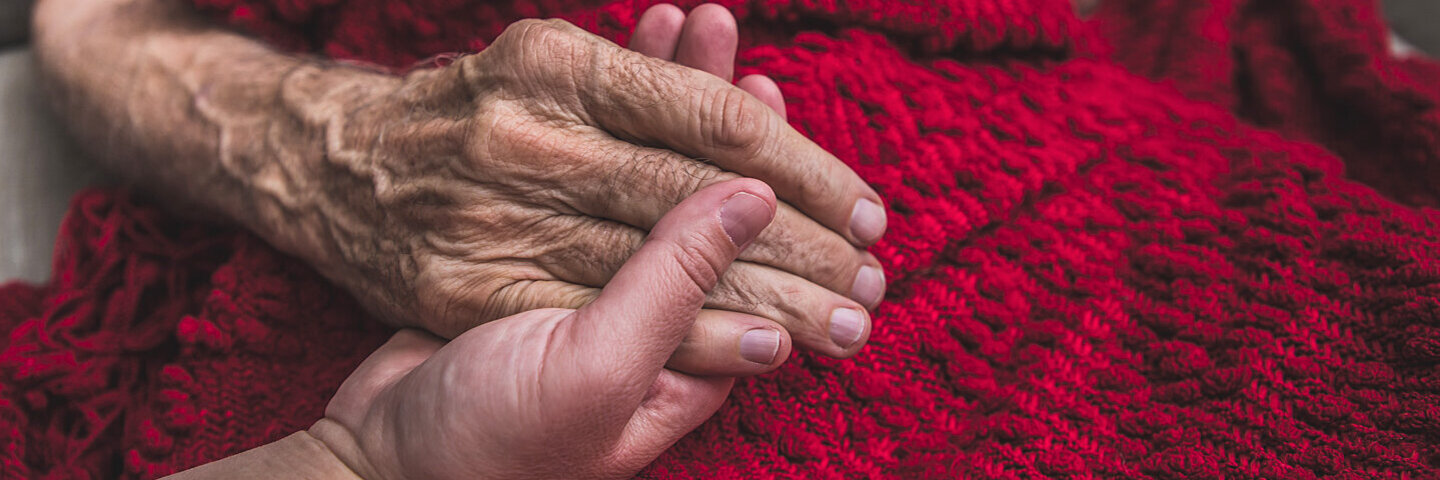 Eine Person hält die Hände einer sterbenden, alten Person mit einer roten Wolldecke.