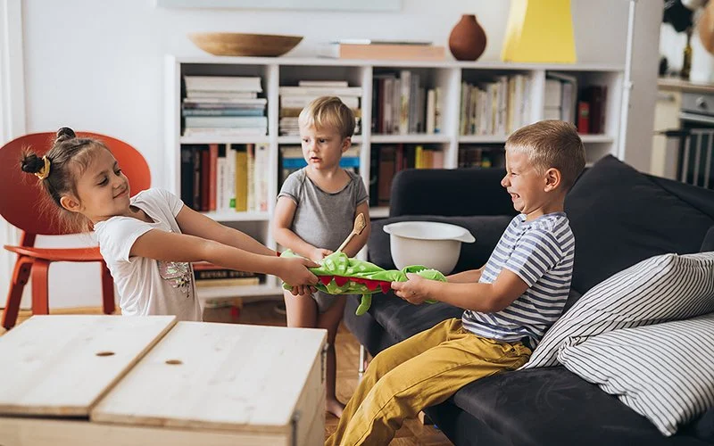 Zwei Jungs und ein Mädchen streiten sich in einem Wohnzimmer um Spielzeug, sie lachen dabei.
