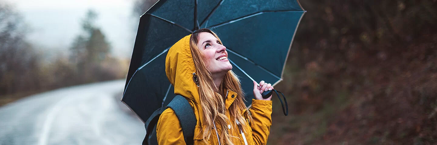 Eine junge Frau steht im Regen, über sich hat sie einen Regenschirm gespannt. Sie schaut zuversichtlich in den Himmel.