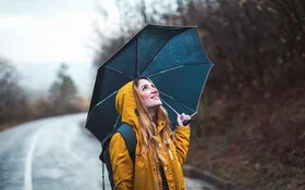 Eine junge Frau steht im Regen, über sich hat sie einen Regenschirm gespannt. Sie schaut zuversichtlich in den Himmel.