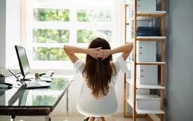 Eine Frau streckt sich mit verschränkten Armen hinter dem Kopf auf Ihrem Büro-Stuhl und guckt dabei aus dem Fenster.