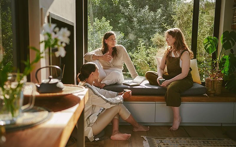 Drei Frauen unterschiedlicher Altersgruppen sitzen in entspannter Atmosphäre vor einem Fenster mit Blick auf die Natur zusammen und unterhalten sich.