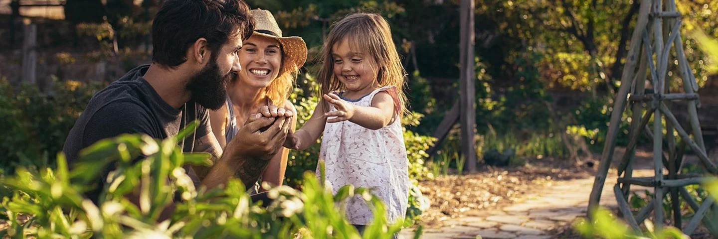 Ein Mann, eine Frau und ein Kind sitzen in einem Garten zwischen grünen Pflanzen. Die Frau trägt einen Strohhut und lächelt, das Kind streckt die Hand aus und lacht.