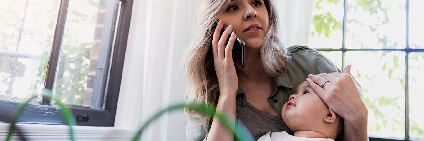 Eine junge Mutter hält die Stirn ihres Kindes und ruft ein Baby-Telefon für Rat an.